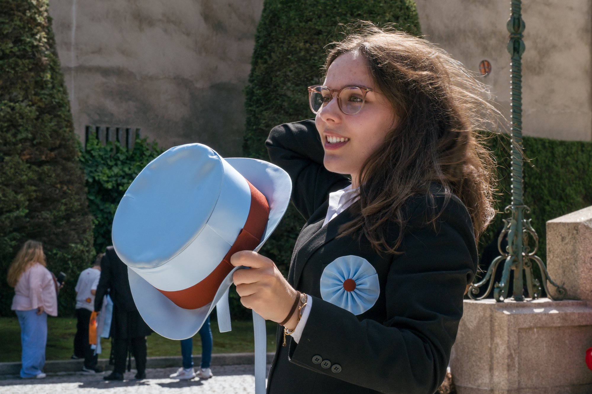 A young woman outdoors holding a ceremonial top hat, hair lifted by the wind, caught mid-sentence and half-smiling.