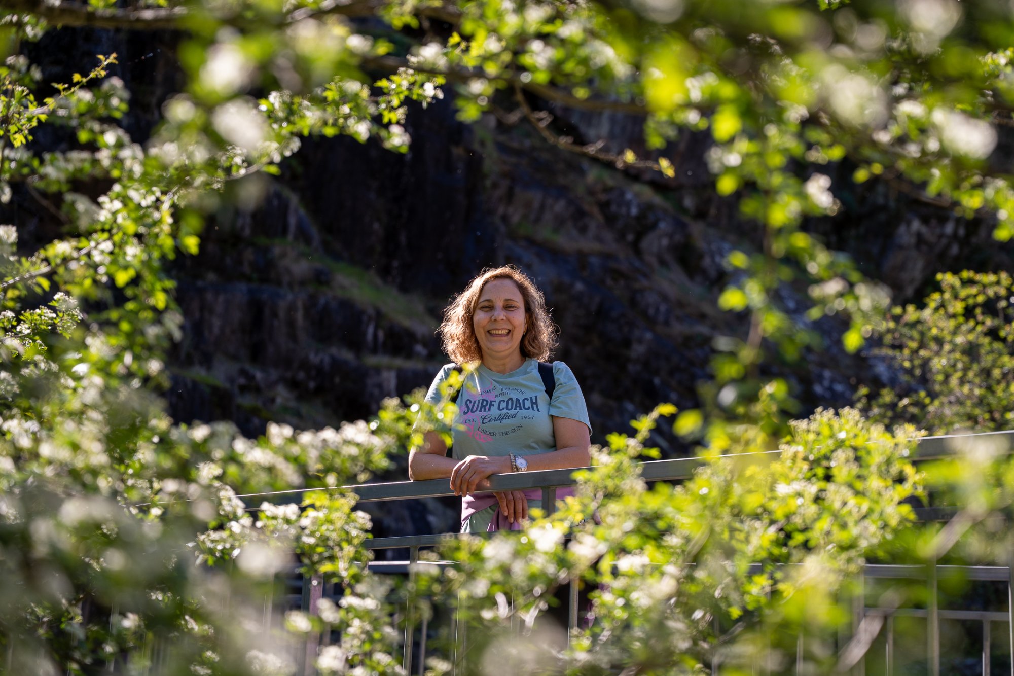 A woman laughing, framed by out-of-focus green spring leaves, sunlight catching her hair as she leans on a railing.